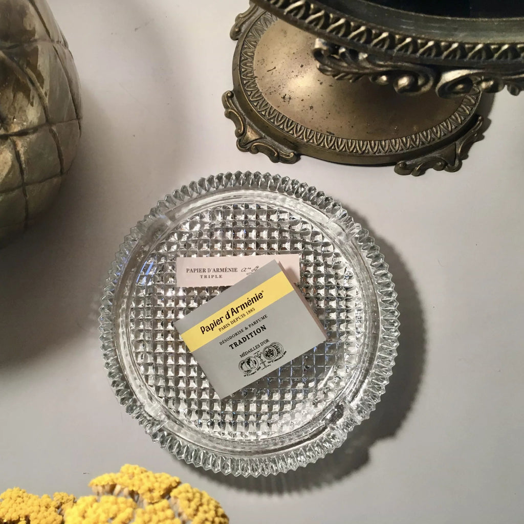 Clear glass tray with a papier d'armenie paper incense on a light grey surface, surrounded by decorative items.