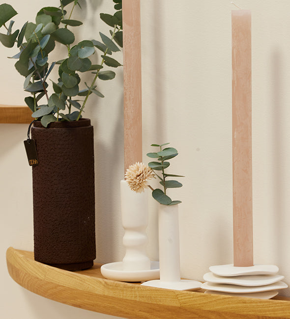 Decorative shelf with a plant in a brown vase, white candle holders, and small decorative items against a light curtain background.
