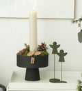 Decorative candle holder with a candle, pine cones, and a small figurine on a white surface.