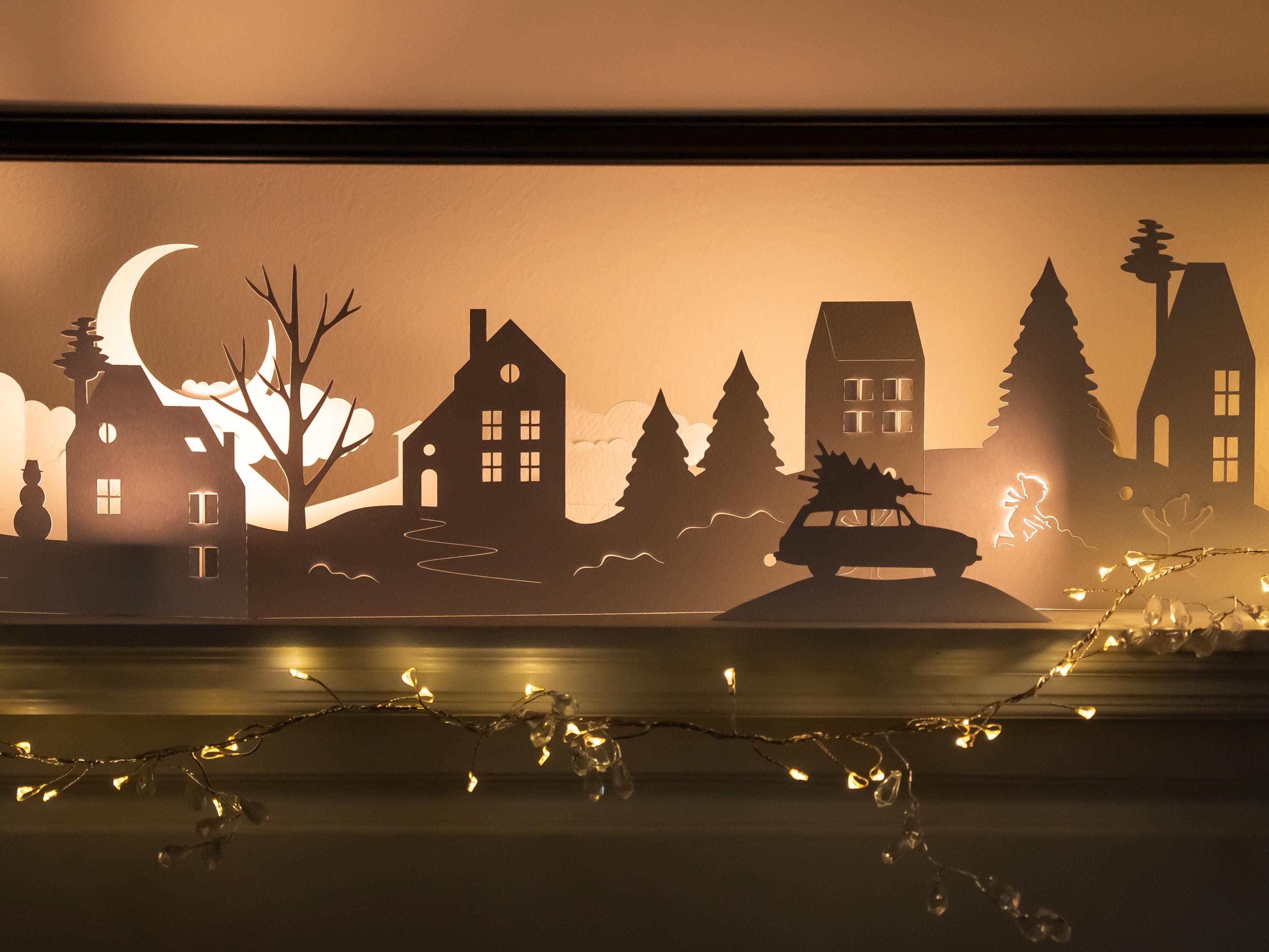 Silhouette scene of houses and trees with a car, illuminated by string lights against a warm background.