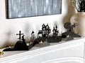 A Halloween-themed tabletop decoration set arranged on a mantel, featuring black silhouettes of a haunted house, cemetery, and other Halloween-related imagery with spaces for LED tealights.